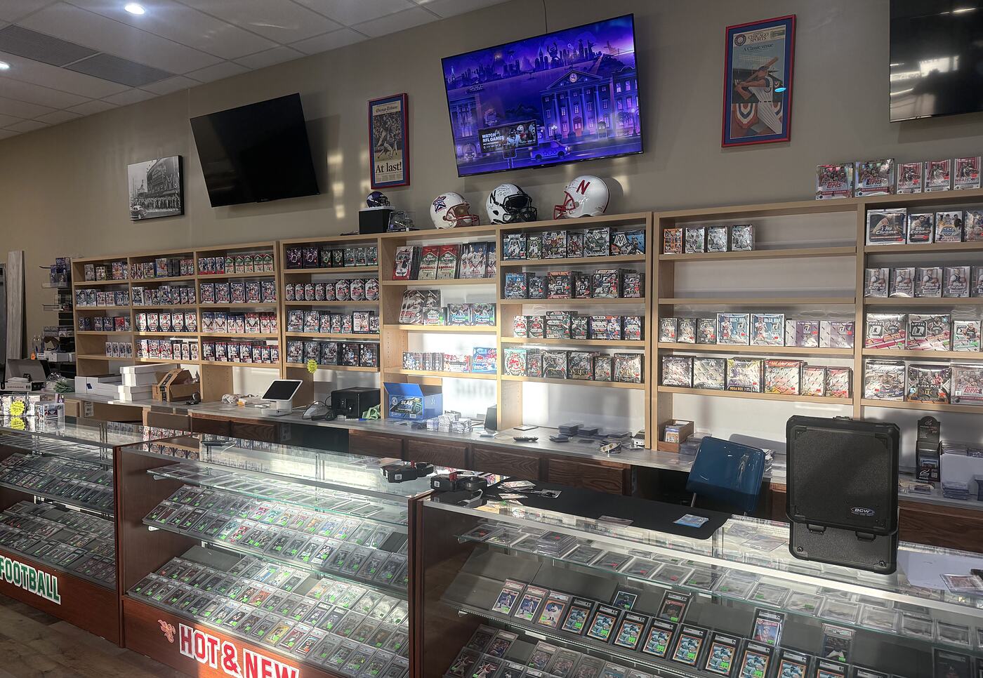 The front counter at Hot Corner in Kearney: register and supplies on the bar, the sealed wax wall and team helmets behind it, and the graded card cases running along the front.