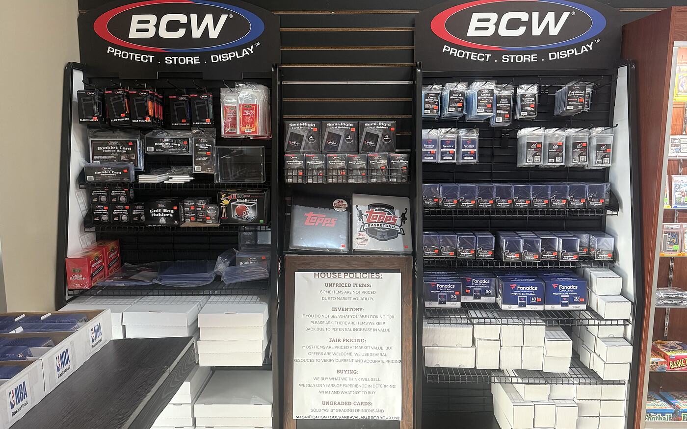 The supplies wall at the Kearney shop: pegged sleeves, top loaders, semi-rigids and one-touch holders beside stacks of white storage boxes and the house policies sign.